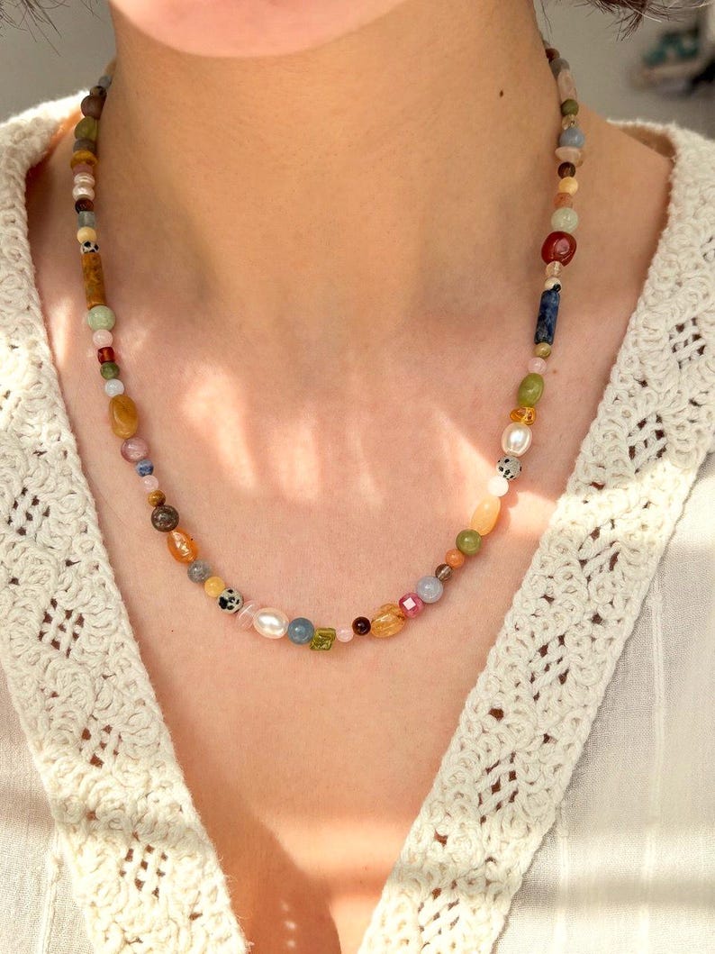 May include: A colorful beaded necklace featuring a variety of natural stone beads in shades of brown, green, yellow, pink, and blue. The necklace is worn around the neck and is a unique piece of jewelry.