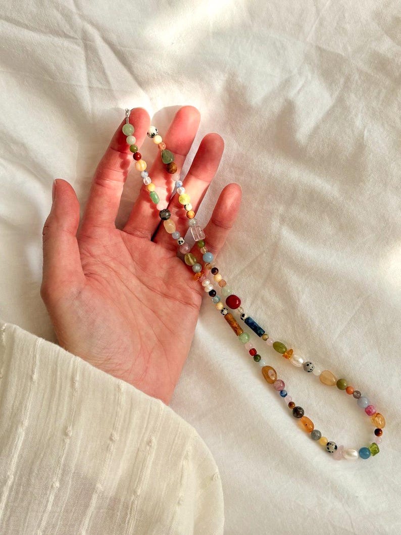 May include: A colorful beaded necklace with a variety of shapes and sizes of beads. The beads are in shades of red, yellow, green, blue, and brown. The necklace is held against a white background.