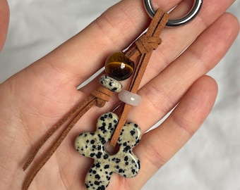 May include: A keychain featuring a silver-toned metal ring attached to brown suede leather cords. The cords are adorned with a brown tiger eye bead, a small dalmatian jasper bead, a pale pink bead, and a dalmatian jasper flower-shaped charm.