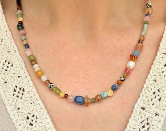 Peut inclure: Un collier de perles colorées avec une variété de pierres précieuses et de perles dans les tons de bleu, vert, rose, jaune et marron. Le collier est enfilé et porté autour du cou.