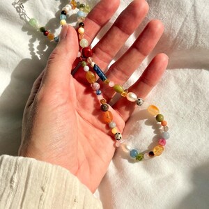 May include: A colorful beaded necklace with a variety of shapes and sizes. The necklace features a mix of gemstones, pearls, and other beads in shades of red, green, yellow, and blue. The necklace is held in a hand.