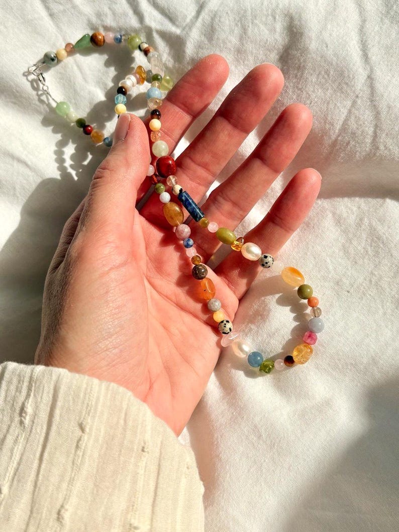 May include: A colorful beaded necklace with a variety of shapes and sizes. The necklace features a mix of gemstones, pearls, and other beads in shades of red, green, yellow, and blue. The necklace is held in a hand.