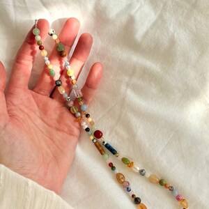 May include: A colorful beaded necklace featuring a variety of gemstones and pearls. The necklace includes beads in shades of red, green, yellow, blue, and brown. The necklace is held against a white background.