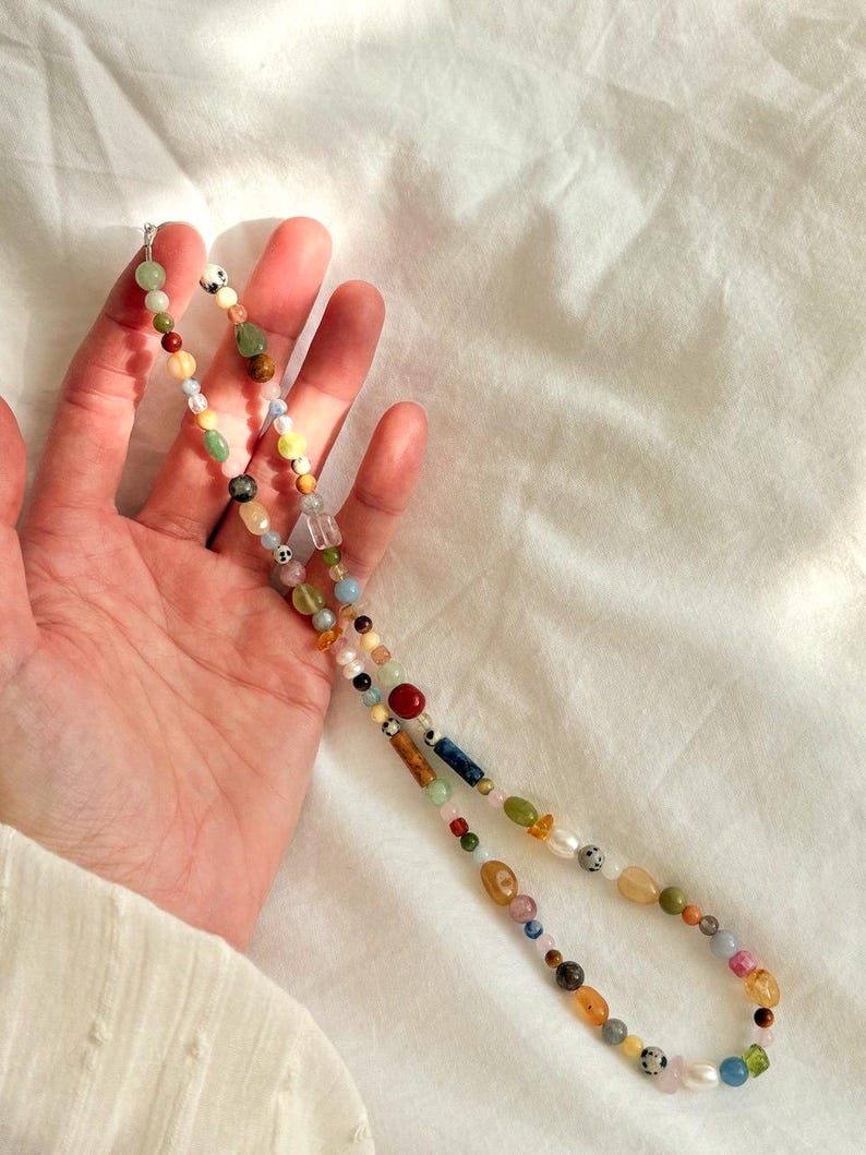 May include: A colorful beaded necklace featuring a variety of gemstones and pearls. The necklace includes beads in shades of red, green, yellow, blue, and brown. The necklace is held against a white background.