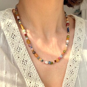May include: A colorful beaded necklace with a variety of shapes and sizes. The necklace features a mix of gemstones, pearls, and other beads in shades of yellow, pink, blue, and green. The necklace is worn around the neck.