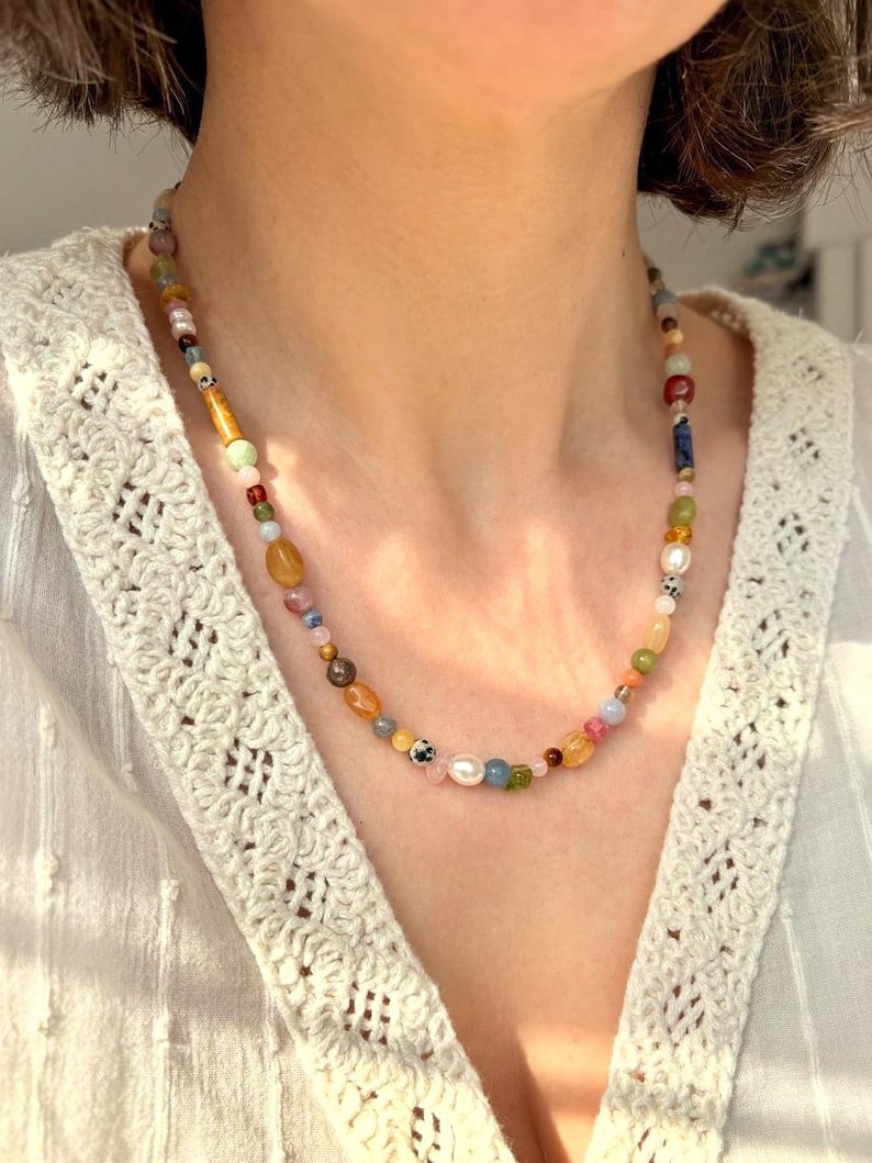 May include: A colorful beaded necklace with a variety of shapes and sizes. The necklace features a mix of gemstones, pearls, and other beads in shades of yellow, pink, blue, and green. The necklace is worn around the neck.