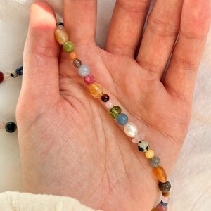 May include: A colorful beaded necklace featuring a variety of gemstones and pearls. The beads are in shades of green, blue, pink, yellow, and orange. The necklace is held in a hand against a white background.