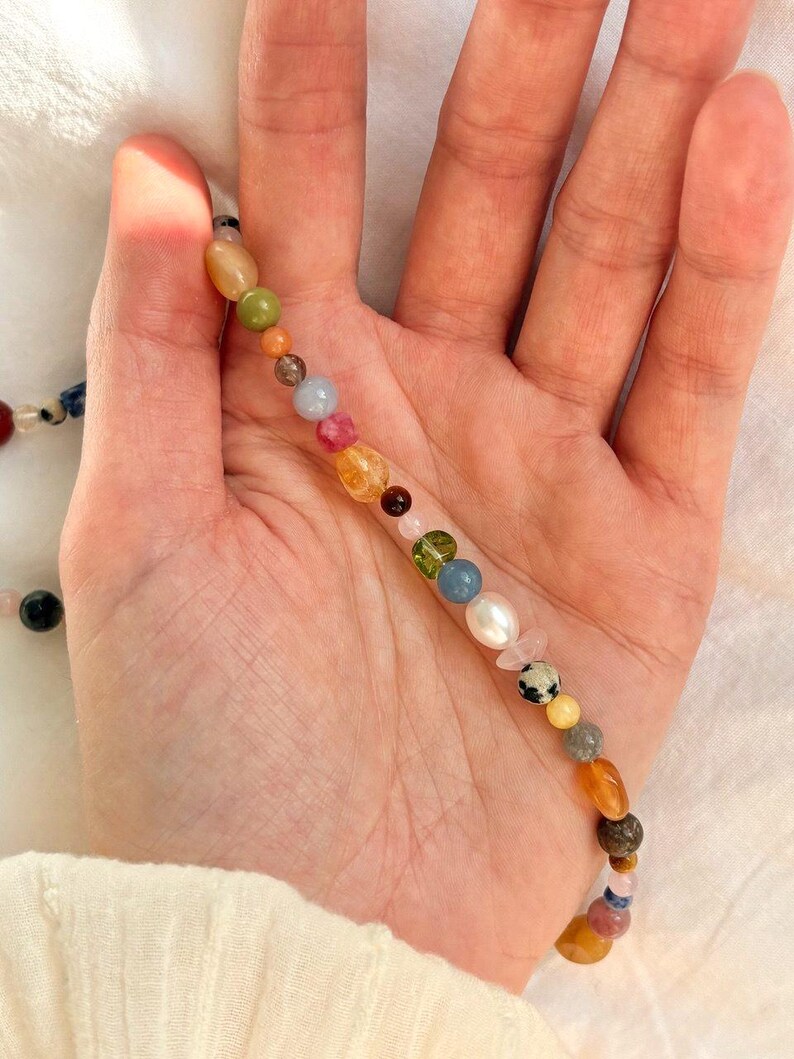 May include: A colorful beaded necklace featuring a variety of gemstones and pearls. The beads are in shades of green, blue, pink, yellow, and orange. The necklace is held in a hand against a white background.