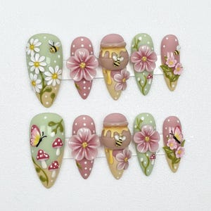 May include: A set of ten almond-shaped false nails with various designs. The nails feature daisies, bees, butterflies, mushrooms, and honey pots with flowers. The color palette includes green, pink, yellow, and white.