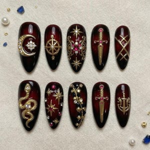 May include: A set of ten long, oval-shaped press-on nails with a dark red to black gradient. Each nail features gold and jewel embellishments, including a crescent moon, compass, stars, swords, and a snake.