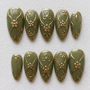 Puede incluir: Uñas postizas verde oliva con diseños florales y de remolinos dorados. Las uñas tienen forma ovalada y presentan intrincados adornos dorados, incluyendo flores y líneas curvas. El conjunto incluye varias tallas.
