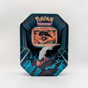 May include: A teal and black Pokémon trading card tin featuring a Paldean Tauros card. The tin has a geometric design and the Pokémon Trading Card Game logo. The card shows a fiery illustration of a creature.