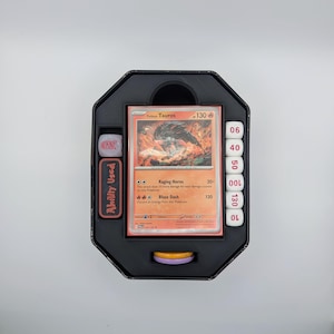 May include: A black, octagonal card game organiser with a Pokémon card featuring a fiery Tauros illustration. Includes dice, damage counters, and a slot for an "Ability Used" marker. The card shows the text "Raging Horns" and "Blaze Dash".