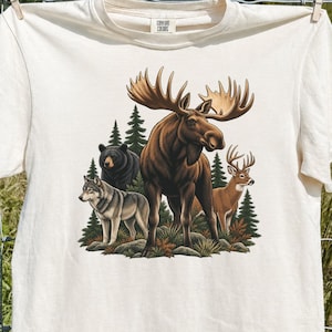 May include: A cream-colored short-sleeve t-shirt featuring a graphic print of a large brown moose standing in the center, surrounded by a black bear, a grey wolf, and a brown deer, all set against a backdrop of green pine trees and foliage.