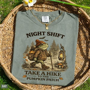 May include: A sage green t-shirt featuring a graphic of a frog wearing a brown beanie and jacket, carrying a backpack with a pumpkin and a lantern. The text reads NIGHT SHIFT TAKE A HIKE TO THE PUMPKIN PATCH. The shirt has a Comfort Colors label.