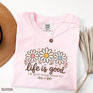 Peut inclure: Un t-shirt rose clair avec un motif de marguerites aux centres roses, jaunes et bleus, et l'inscription "life is good". Un collier dor&eacute; avec un pendentif est pos&eacute; sur le t-shirt. Le mot "Blossom" est en bas &agrave; gauche.