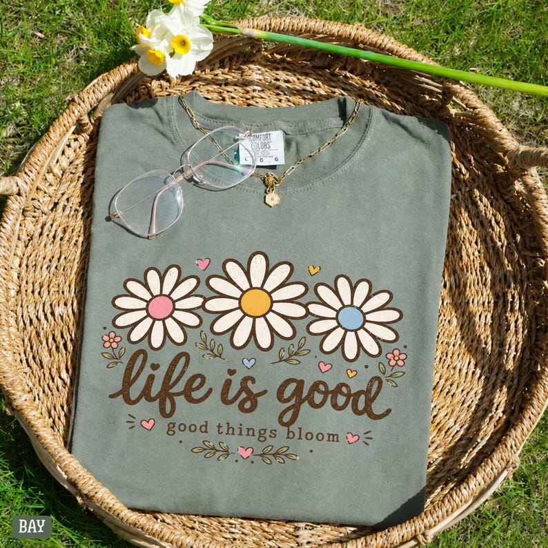 Peut inclure: T-shirt vert sauge avec un motif de marguerites et le texte "life is good, good things bloom." Le t-shirt est pr&eacute;sent&eacute; dans un panier en osier avec un collier en or et des lunettes. Une branche de jonquilles est en arri&egrave;re-plan.