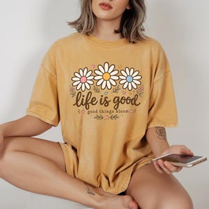 Peut inclure: T-shirt jaune moutarde avec un motif de marguerites et le texte "life is good good things bloom". Le motif pr&eacute;sente trois marguerites avec des centres roses, blancs et bleus. Le t-shirt est de coupe ample.
