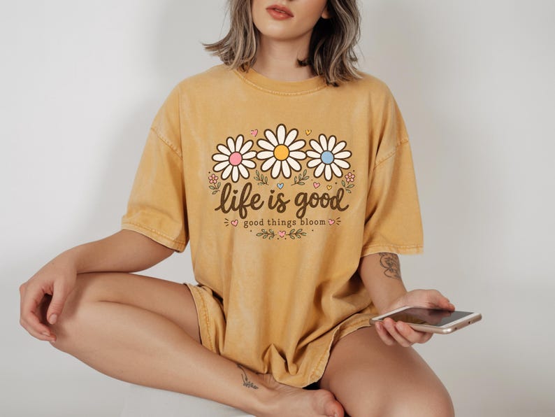 Peut inclure: T-shirt jaune moutarde avec un motif de marguerites et le texte "life is good good things bloom". Le motif pr&eacute;sente trois marguerites avec des centres roses, blancs et bleus. Le t-shirt est de coupe ample.