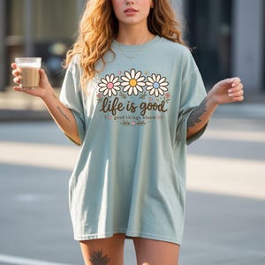 Peut inclure: T-shirt bleu clair avec un motif de marguerites et le texte "life is good good things bloom". Le motif de marguerite pr&eacute;sente des p&eacute;tales blancs, roses et jaunes. Le t-shirt a des manches courtes et une coupe ample.