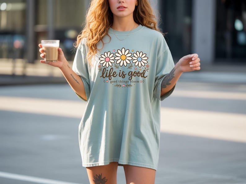 Peut inclure: T-shirt bleu clair avec un motif de marguerites et le texte "life is good good things bloom". Le motif de marguerite pr&eacute;sente des p&eacute;tales blancs, roses et jaunes. Le t-shirt a des manches courtes et une coupe ample.
