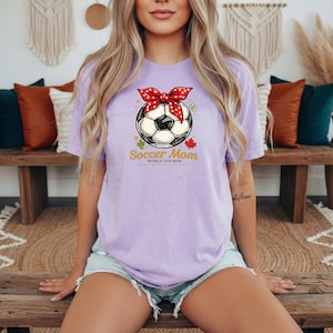 May include: A light purple t-shirt featuring a soccer ball design with a red bow and the text "Soccer Mom World Cup 2026". The graphic includes a soccer ball, maple leaf, and flower. The shirt is made of a soft material.