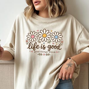 Peut inclure: T-shirt beige avec un motif floral. Le motif pr&eacute;sente trois marguerites roses, jaunes et bleues, avec les mots "life is good" et "good things bloom" en &eacute;criture marron. Le t-shirt est de coupe ample.