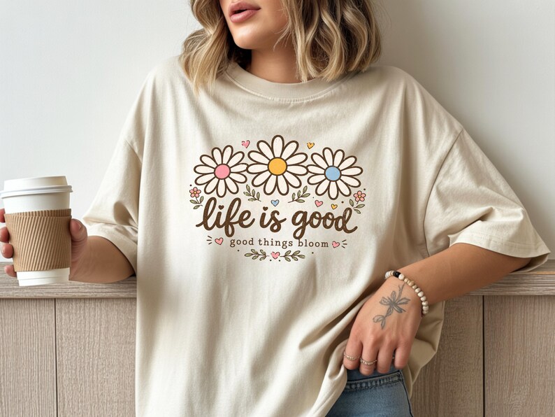 Peut inclure: T-shirt beige avec un motif floral. Le motif pr&eacute;sente trois marguerites roses, jaunes et bleues, avec les mots "life is good" et "good things bloom" en &eacute;criture marron. Le t-shirt est de coupe ample.