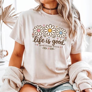 Peut inclure: T-shirt beige avec un motif floral de trois marguerites roses, jaunes et bleues. Le texte "life is good, good things bloom" est &eacute;crit en &eacute;criture cursive. Le t-shirt est &agrave; manches courtes.
