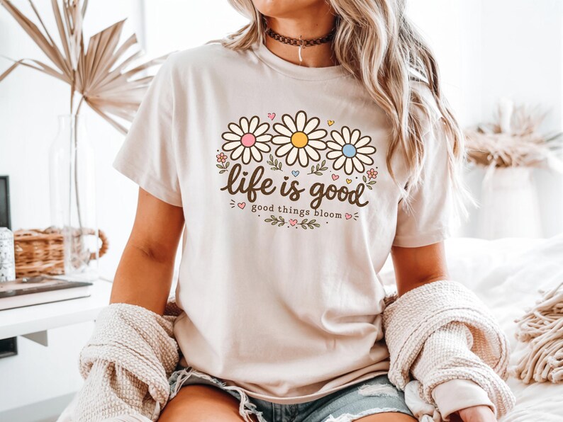 Peut inclure: T-shirt beige avec un motif floral de trois marguerites roses, jaunes et bleues. Le texte "life is good, good things bloom" est &eacute;crit en &eacute;criture cursive. Le t-shirt est &agrave; manches courtes.