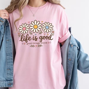 Peut inclure: Un t-shirt rose clair avec l'inscription "life is good" et "good things bloom" en script marron. Le t-shirt pr&eacute;sente trois fleurs de marguerite avec des centres roses, jaunes et bleus. Le t-shirt est port&eacute; avec une veste en jean bleue.