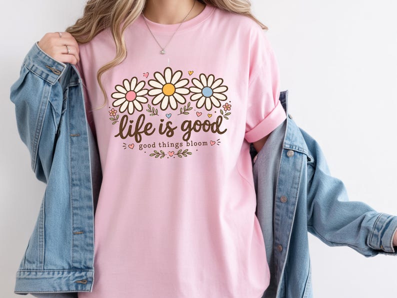 Peut inclure: Un t-shirt rose clair avec l'inscription "life is good" et "good things bloom" en script marron. Le t-shirt pr&eacute;sente trois fleurs de marguerite avec des centres roses, jaunes et bleus. Le t-shirt est port&eacute; avec une veste en jean bleue.