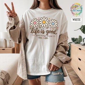 Peut inclure: T-shirt blanc avec un motif de marguerites aux c&oelig;urs roses, jaunes et bleus. Le texte "life is good" est au-dessus des fleurs, et "good things bloom" en dessous. V&ecirc;tement d&eacute;contract&eacute;.