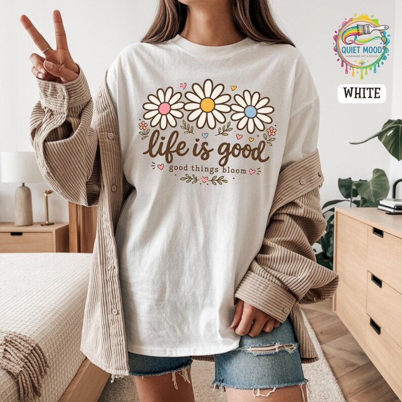 Peut inclure: T-shirt blanc avec un motif de marguerites aux c&oelig;urs roses, jaunes et bleus. Le texte "life is good" est au-dessus des fleurs, et "good things bloom" en dessous. V&ecirc;tement d&eacute;contract&eacute;.