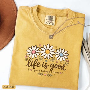 Peut inclure: T-shirt moutarde avec un motif de marguerites et l'inscription "life is good" et "good things bloom". Le t-shirt a un col rond et des manches courtes. Un collier argent&eacute; avec un pendentif est pos&eacute; sur le t-shirt.