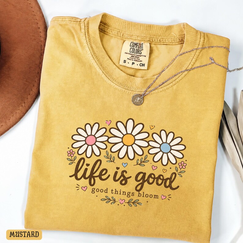 Peut inclure: T-shirt moutarde avec un motif de marguerites et l'inscription "life is good" et "good things bloom". Le t-shirt a un col rond et des manches courtes. Un collier argent&eacute; avec un pendentif est pos&eacute; sur le t-shirt.