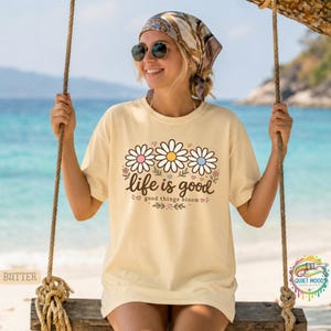 Peut inclure: T-shirt jaune clair avec l'inscription "life is good" et trois fleurs de marguerite roses, jaunes et bleues. Le t-shirt a une coupe d&eacute;contract&eacute;e, id&eacute;al pour un usage quotidien.