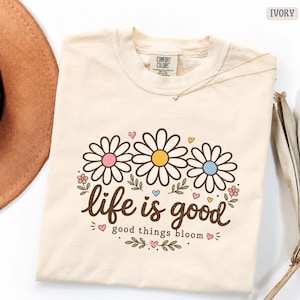 Peut inclure: T-shirt ivoire avec un motif de marguerites. Le motif pr&eacute;sente trois marguerites avec des centres roses, jaunes et bleus, accompagn&eacute;es des mots "life is good" et "good things bloom". Un collier en or est drap&eacute; sur le t-shirt.