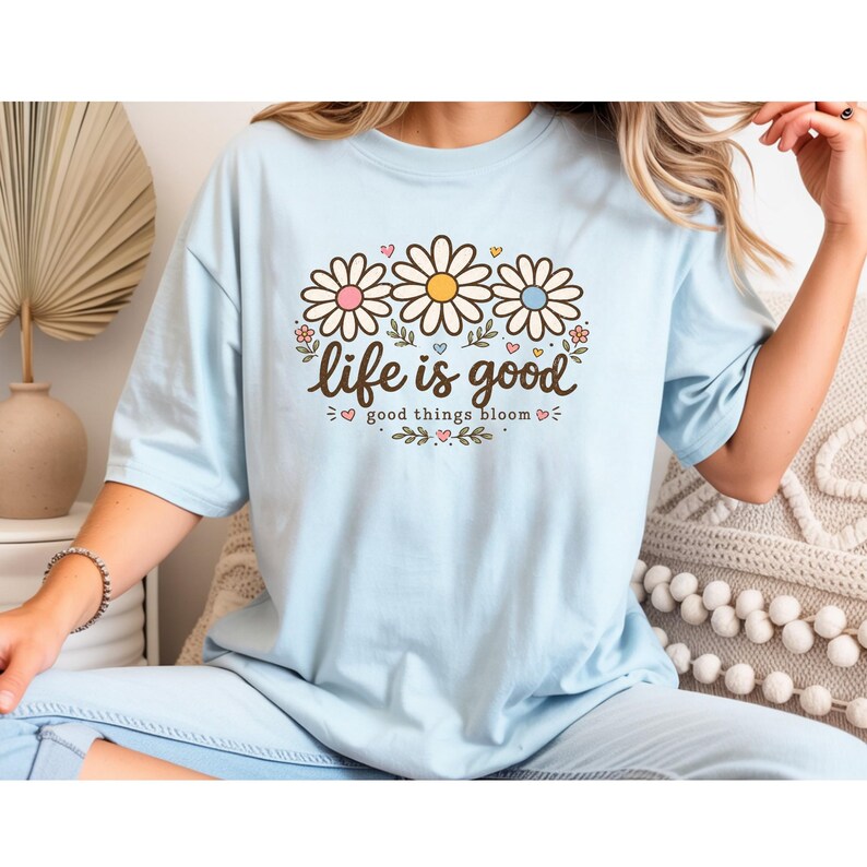 Peut inclure: T-shirt bleu clair avec un motif floral de marguerites. Le motif pr&eacute;sente trois marguerites avec des centres roses, jaunes et bleus, et le texte "life is good" avec "good things bloom" en dessous. V&ecirc;tement d&eacute;contract&eacute;.
