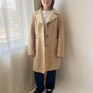 May include: A tan knee-length coat featuring a cream-colored faux shearling collar and lapels. The coat has a button-front closure with three visible buttons, long sleeves, and two vertical side pockets. It is worn over a white shirt and blue jeans.