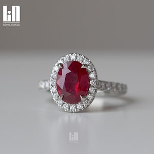 Può includere: Anello in argento con un grande rubino ovale circondato da un alone di diamanti più piccoli. La fascia dell'anello è anch'essa ornata di diamanti. Il logo Sons Jewels è visibile.