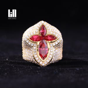 May include: A gold-toned ring with a large, red gemstone in a cross-like design. The ring is encrusted with small, clear gemstones. The brand name "SONS JEWELS" is visible in the upper left corner.