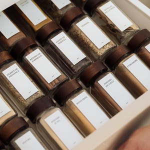 May include: A close-up of a drawer containing glass spice jars with dark wooden lids and white labels. Spices include cinnamon sticks, chilli powder, and coriander. The jars are neatly arranged.