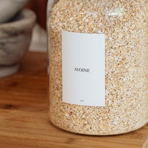 May include: A glass jar filled with rolled oats, labelled "AVOINE". The jar is on a wooden surface. A marble mortar and pestle are in the background. The oats are a light tan colour. The label is white with black text.