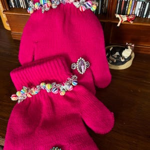 May include: A pair of bright pink mittens adorned with colorful beaded accents and sparkling embellishments. One mitten features a large, oval-shaped ring with a purple stone, while the other has a round, blue and silver brooch.