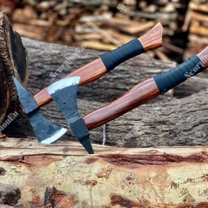 May include: Two axes with wooden handles and black blades are displayed. The handles are a rich brown color, wrapped with black material near the head. The axes are resting on a wooden surface, showcasing their craftsmanship.