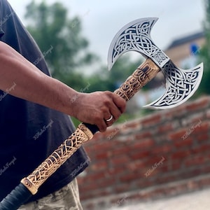 Puede incluir: Un hacha de batalla de doble filo con una gran cabeza de metal plateado y grabados intrincados de nudos celtas negros. El mango largo está hecho de madera clara con patrones tallados detallados y una empuñadura negra en la base.