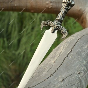 May include: A decorative dagger with a silver-colored handle featuring skulls and a crown. The blade is a bright, polished silver. The dagger is resting on a weathered, gray tree branch, with a blurred green background.
