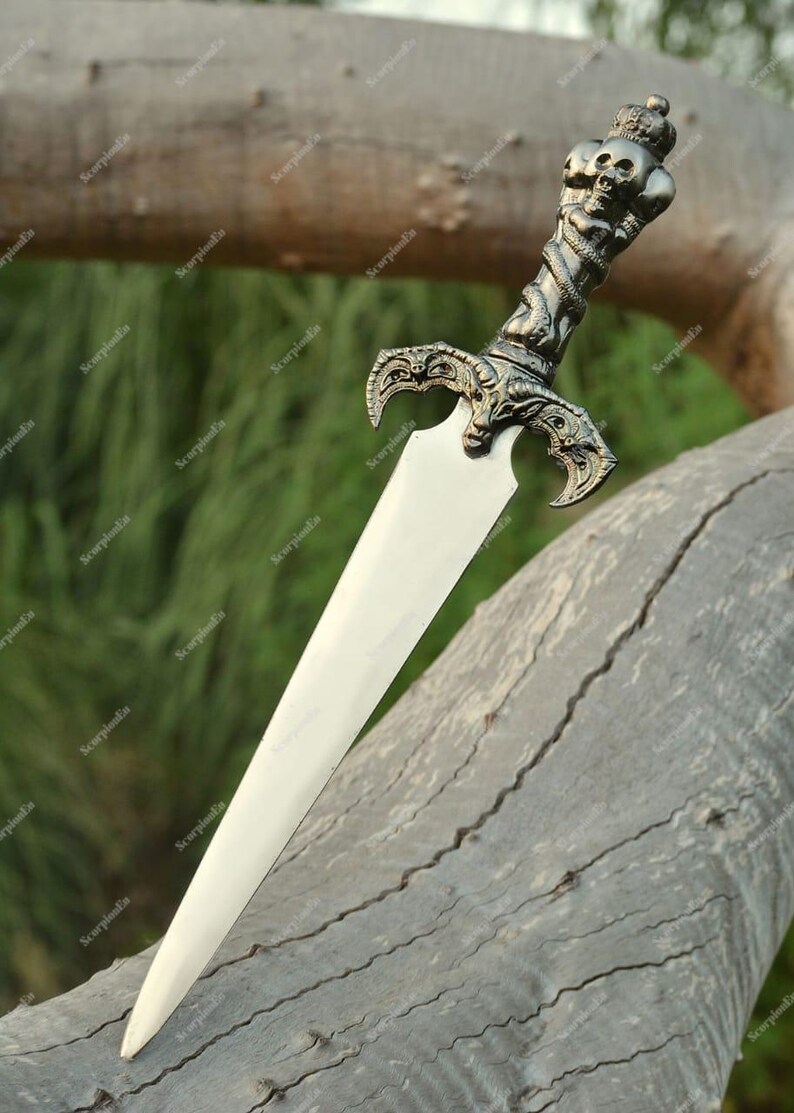 May include: A decorative dagger with a silver-colored handle featuring skulls and a crown. The blade is a bright, polished silver. The dagger is resting on a weathered, gray tree branch, with a blurred green background.