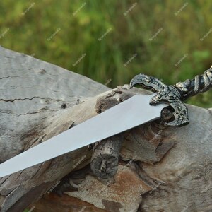 May include: A decorative dagger with a silver blade and an ornate handle. The handle features a detailed design with a dark metallic finish. The dagger is resting on a weathered wooden log, with a blurred green background.